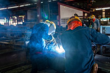 Kaynakçı, Flux Cored Arc Welding (FCAW) işlemiyle bir çelik yapı çalışması yapıyor ve endüstriyel fabrikada güvenlik için kişisel koruyucu ekipman (PPE) ile uygun şekilde giyiniyor.