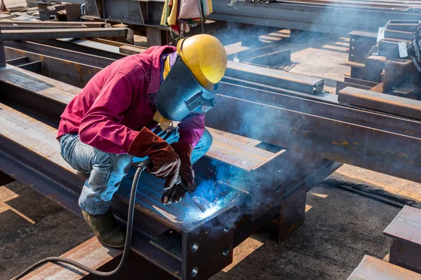 Kaynakçı, Flux Cored Arc Welding (FCAW) işlemiyle bir çelik yapı çalışması yapıyor ve endüstriyel fabrikada güvenlik için kişisel koruyucu ekipman (PPE) ile uygun şekilde giyiniyor.