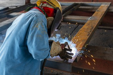 Kaynakçı, Flux Cored Arc Welding (FCAW) işlemiyle bir çelik yapı çalışması yapıyor ve endüstriyel fabrikada güvenlik için kişisel koruyucu ekipman (PPE) ile uygun şekilde giyiniyor.