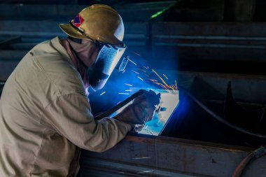 Kaynakçı, Flux Cored Arc Welding (FCAW) işlemiyle bir çelik yapı çalışması yapıyor ve endüstriyel fabrikada güvenlik için kişisel koruyucu ekipman (PPE) ile uygun şekilde giyiniyor.