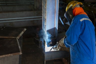 Kaynakçı, Flux Cored Arc Welding (FCAW) işlemiyle bir çelik yapı çalışması yapıyor ve endüstriyel fabrikada güvenlik için kişisel koruyucu ekipman (PPE) ile uygun şekilde giyiniyor.