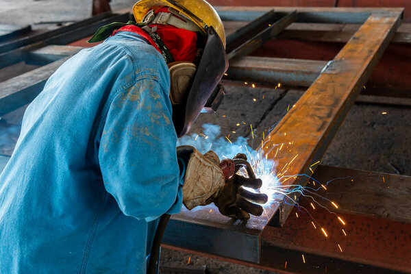 Сварщик сваривает стальную конструкцию по технологии Flux Cored Arc Welding (FAW) и надевает средства индивидуальной защиты (СИЗ) для обеспечения безопасности на промышленном предприятии.