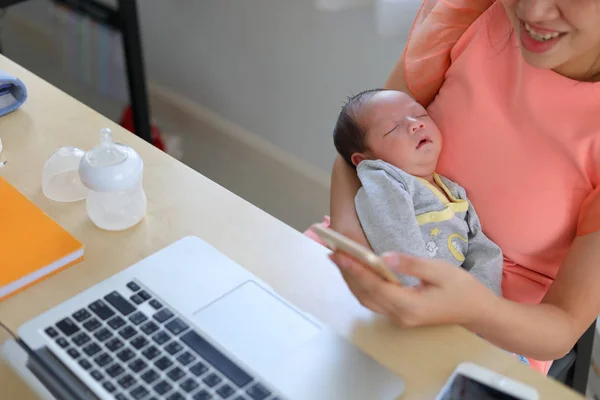 Mãe jogando telefone celular em casa de negócios escritório com bebê bonito recém-nascido dormindo — Fotografia de Stock
