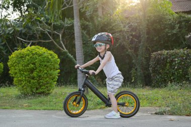 Mutlu şirin çocuk güneş gözlüğü ve kask takıyor ve sabah denge bisikleti egzersizi yapıyor.
