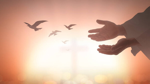 Концепция пасхального воскресенья: силуэт человека открывает две пустые руки с поднятыми ладонями и птицами, пролетающими над размытым крестом на церковном фоне
