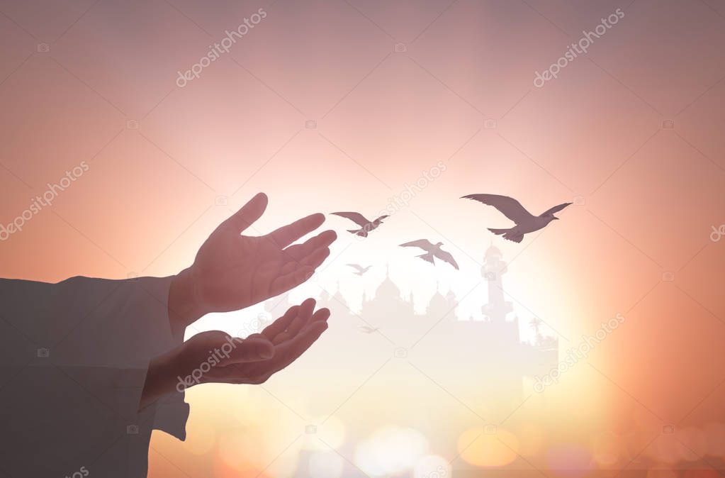 Muslim man open empty hands with palms up with birds flying over blurred mosque on sunset background.