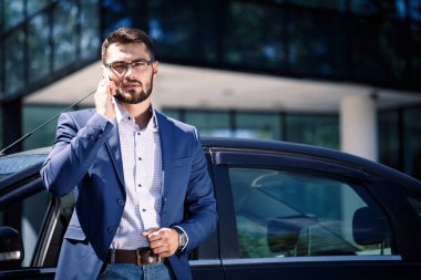 İş adamı arabanın yanında duruyor ve telefonla konuşuyor. Arka plandaki ofis binası.