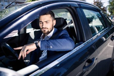 Genç adam arabada oturuyor ve tedirgin gözlerle saate bakıyor. 