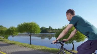 Genç bir adam güneşli bir günde bir nehir boyunca bisiklet sürüyor. Buharlı çekim.
