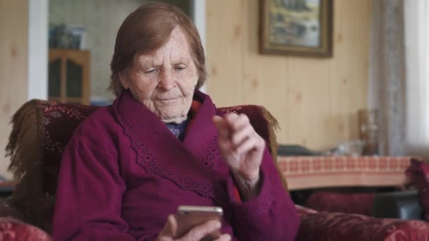 年配の女性がビデオ通話用の携帯電話を持っている 彼女は携帯電話の画面を見て笑顔で手を振って — ストック動画
