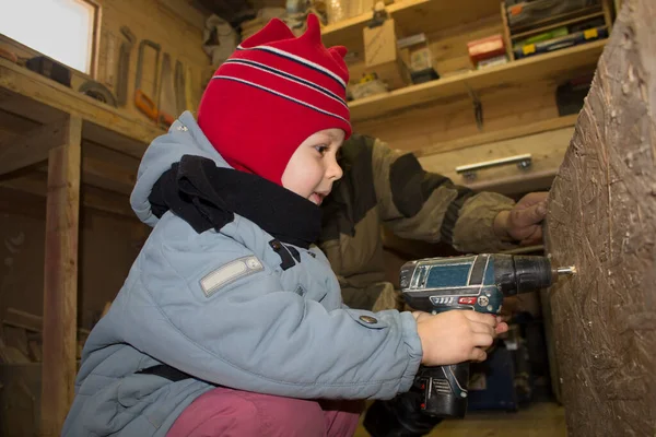 Oğlan tornavidayı ahşap bir duvara deliyor. Kalın giysiler içinde. Marangoz dükkanında.