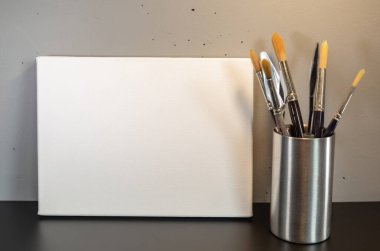 Blank white canvas standing in a landscape form and brusches in a metal can on a black  surface on a gray background.
