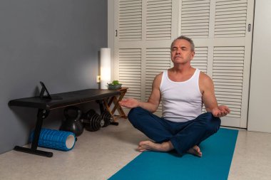 Beyaz atletli ve mavi eşofmanlı orta yaşlı beyaz bir adam evdeki halıda yoga yapıyor..
