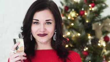 Güzel kız içki şampanya ve gülümseyen, yeni yıl için Noel ağacının yanında duruyor. Çok güzel ve çekici bir kız. Yakın çekim.