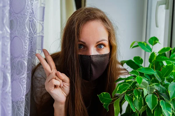 Pencerenin yanındaki pencerenin pervazında koruyucu maskeli bir kız. Gizli virüs salgınına karşı korunma amaçlı. Tedavi, Önleme, Tecrit