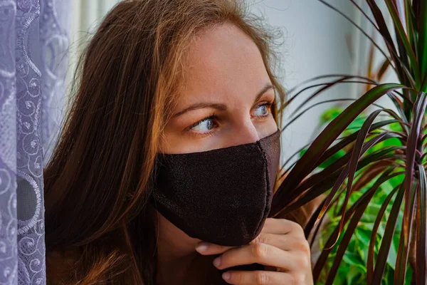 Pencerenin yanındaki pencerenin pervazında koruyucu maskeli bir kız. Gizli virüs salgınına karşı korunma amaçlı. Tedavi, Önleme, Tecrit