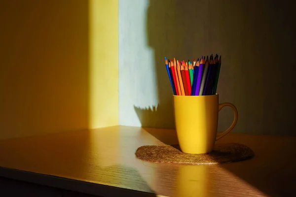 Colored pencils stand in a yellow mug on a table in a ray of sunlight, around a shadow. Office for the student on the desk. The child will draw pictures with a pencil