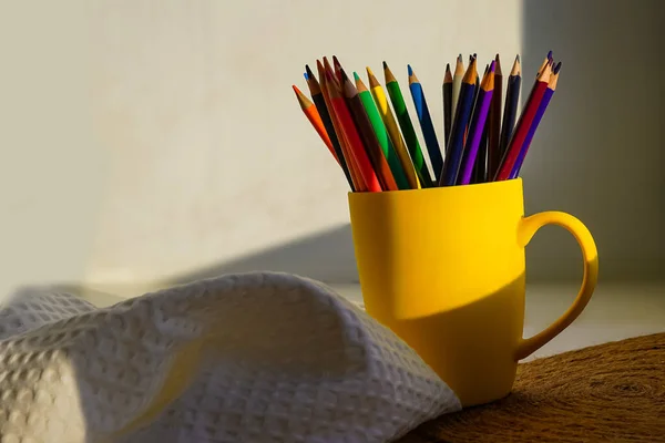 Renkli kalemler sarı bir fincanda duruyor. Masanın üzerinde, güneş ışığı altında, bir gölgenin etrafında. Masadaki öğrenci için ofis. Çocuk kalemle resim çizecek.