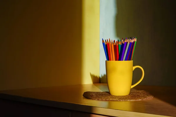 Colored pencils stand in a yellow mug on a table in a ray of sunlight, around a shadow. Office for the student on the desk. The child will draw pictures with a pencil