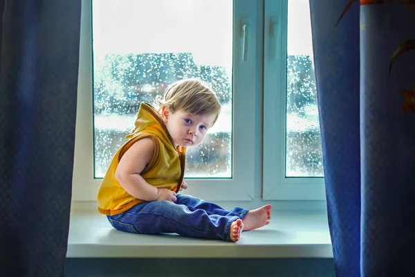 A little boy sits on a window with blue curtains, the bright sun shines from behind, back light, raindrops on the glass