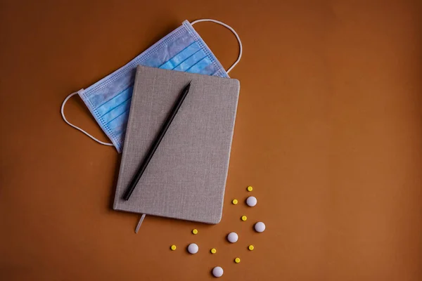 Not defteri ve yuvarlak haplar beyaz ve sarı ve siyah kalem ve kahverengi arka planda mavi koruyucu maske.