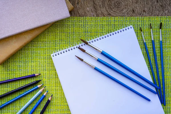 Beyaz bir albümde boya için fırçalar vardır. Kalemlerin ve defterlerin yanında. Gün Işığı Yeşil arka planı. Yaratıcı bir insan için elverişli bir iş yeri. Bir sanatçı için..