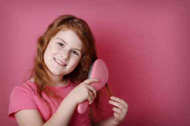 Kızıl saçlı kıvırcık kız saçını tarıyor. Pembe arka planda gülümseyen küçük kız. Boşluğu kopyala.
