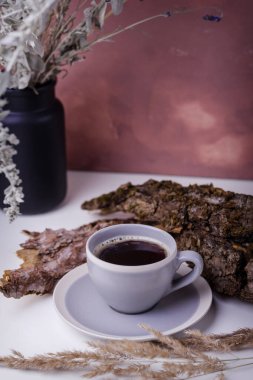 Masadaki bir fincan kahve. Etrafı ağaç kabuğundan dekorla ve kurumuş çiçeklerle dolu vazoyla çevrili. Dekoratif duvar arkaplanı