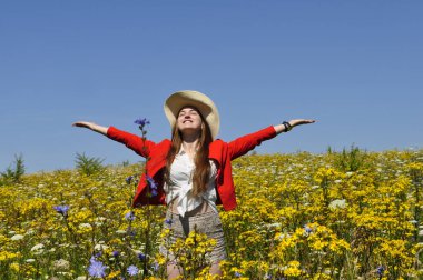 Parlak güneşli bir günde kırmızı ceketli mutlu genç kız çiçek tarlasında duruyor ve ellerini gökyüzüne kaldırıyor.