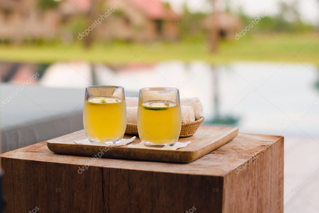 Two yellow lemonade glasses on a wooden tray and wood block next to a swimming pool in boutique hotel resort in Cambodia.