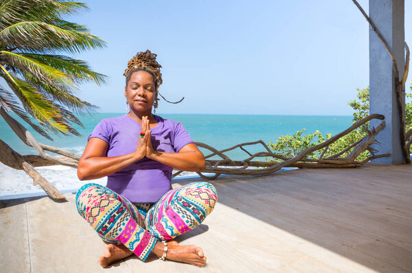 Beautiful Black, African, or Carribean woman practicing wellness and yoga poses at resort or hotel outdoors overlooking gorgeous beach and ocean.