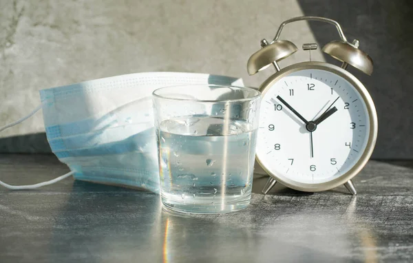 glass of water, alarm clock and medical mask on a gray background