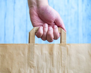 İnsan eli mavi arka planda bir kese kağıdını elinde tutar. Kraft çantası elini tutuyor. Ekolojik kağıt alışveriş torbası adamın elinde. Boşluğu kopyala