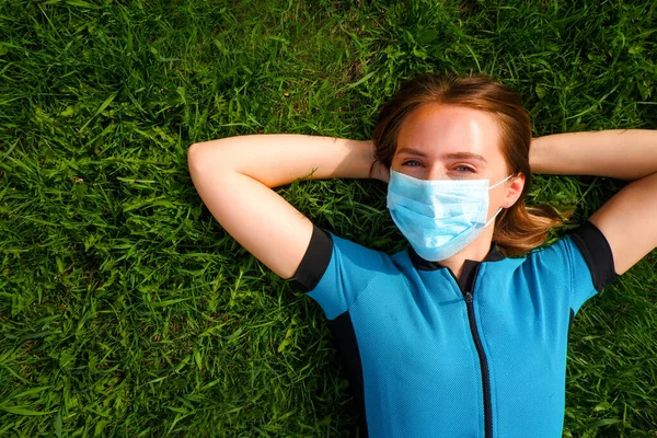 Avrupa görünümlü koruyucu tıbbi maske takmış genç bir kadın parktaki yeşil çimlerin üzerinde karantina sırasında yatıyor. Bahar Yazı. Kız çerçeveye bakıyor..