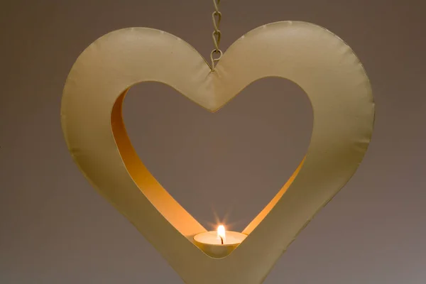 Burning tealight candle in a heart shaped metal candle holder hanging in front of a graduated plain background