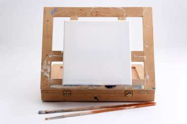 İyi kullanılmış bir ressamın portatif sehpası ve boş bir tuval ve sanat eserinin başlamasını bekleyen iki fırçayla bembeyaz bir tablonun üzerinde duruyor.