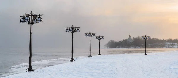 Besneeuwde dijk langs de mistige rivier met lantaarns op het mistige sunrise - winterlandschap. — Stockfoto