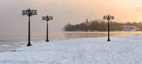 Besneeuwde dijk langs de mistige rivier met lantaarns op het mistige sunrise - winterlandschap. II — Stockfoto