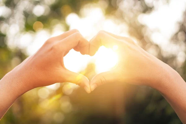 Weibliche Hände Herzform auf Natur Bokeh Sonnenlicht Fackel und verschwimmen Blatt abstrakten Hintergrund. — Stockfoto