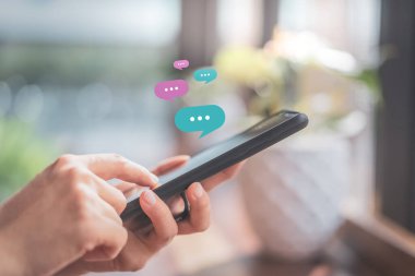 Kadın eli, kafenin soyut arka planında sohbet ikonu grafiği olan akıllı telefon kullanıyor. Teknoloji sektörü ve serbest meslek kavramı. Klasik ton filtre efekti renk biçimi.