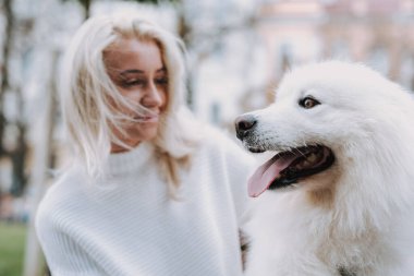 Genç, gülümseyen sarışın kadın parkta beyaz tüylü köpeğini gezdiriyor. Sahip ve evcil hayvan