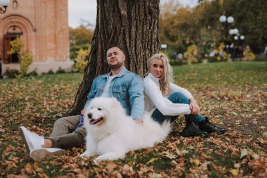 Genç bir kadın ve beyaz tüylü köpekli bir adam sonbahar parkında ağacın altında oturuyorlar. Yeşil çimlerde sarı yapraklar