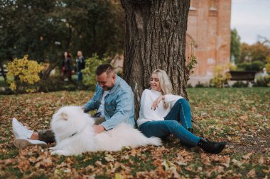 Genç bayan ve beyaz tüylü köpeği olan erkek arkadaşı sonbahar parkında ağacın altında oturuyorlar.