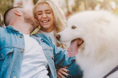 Mutlu genç bir kadınla beyaz tüylü köpekli bir adam, sonbahar parkında ağacın altında öpüşüyor.