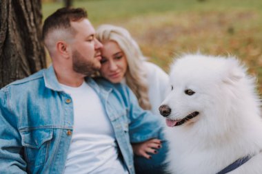 Mutlu dişi ve erkek, beyaz tüylü köpek yavrusuyla sonbahar parkında ağacın altında otururken doğanın ve temiz havanın tadını çıkarıyor.