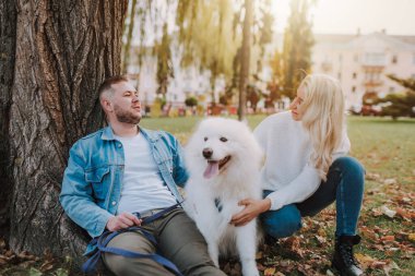 Mutlu genç kadın ve adam, güz parkında ağacın altında otururken beyaz tüylü, oynak köpeğe sarılıyorlar.