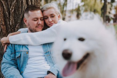 Mutlu koca ve karısı, sonbahar parkında büyük bir ağacın yanında, beyaz tüylü, oynak bir köpekle poz veriyorlar.