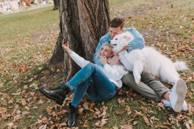 Parkta oturmuş akıllı telefondan fotoğraf çeken, köpekle gülümseyen genç çift.
