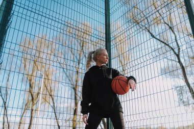 Siyah spor kıyafetli bir kadının elinde basketbol topuyla başka yere baktığı düşük açı.