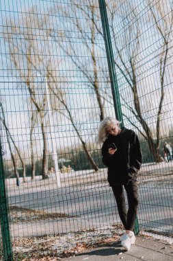 Metal şebekenin yanında duran güzel bir bayan akıllı telefonu tutuyor ve dışarıda yazı yazıyor.